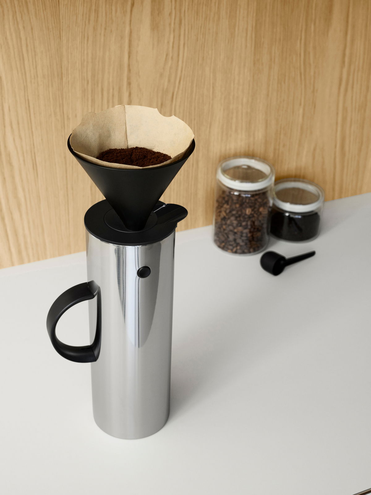 Stelton Filterhalter auf einer silbernen Isolierkanne, seitliche Perspektive, mit Kaffeefilter und Kaffeepulver, im Hintergrund zwei Gläser mit Kaffeebohnen und Kaffeepulver.