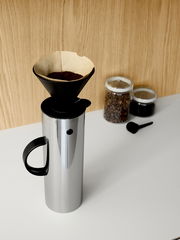 Stelton Filterhalter auf einer silbernen Isolierkanne, seitliche Perspektive, mit Kaffeefilter und Kaffeepulver, im Hintergrund zwei Gläser mit Kaffeebohnen und Kaffeepulver.