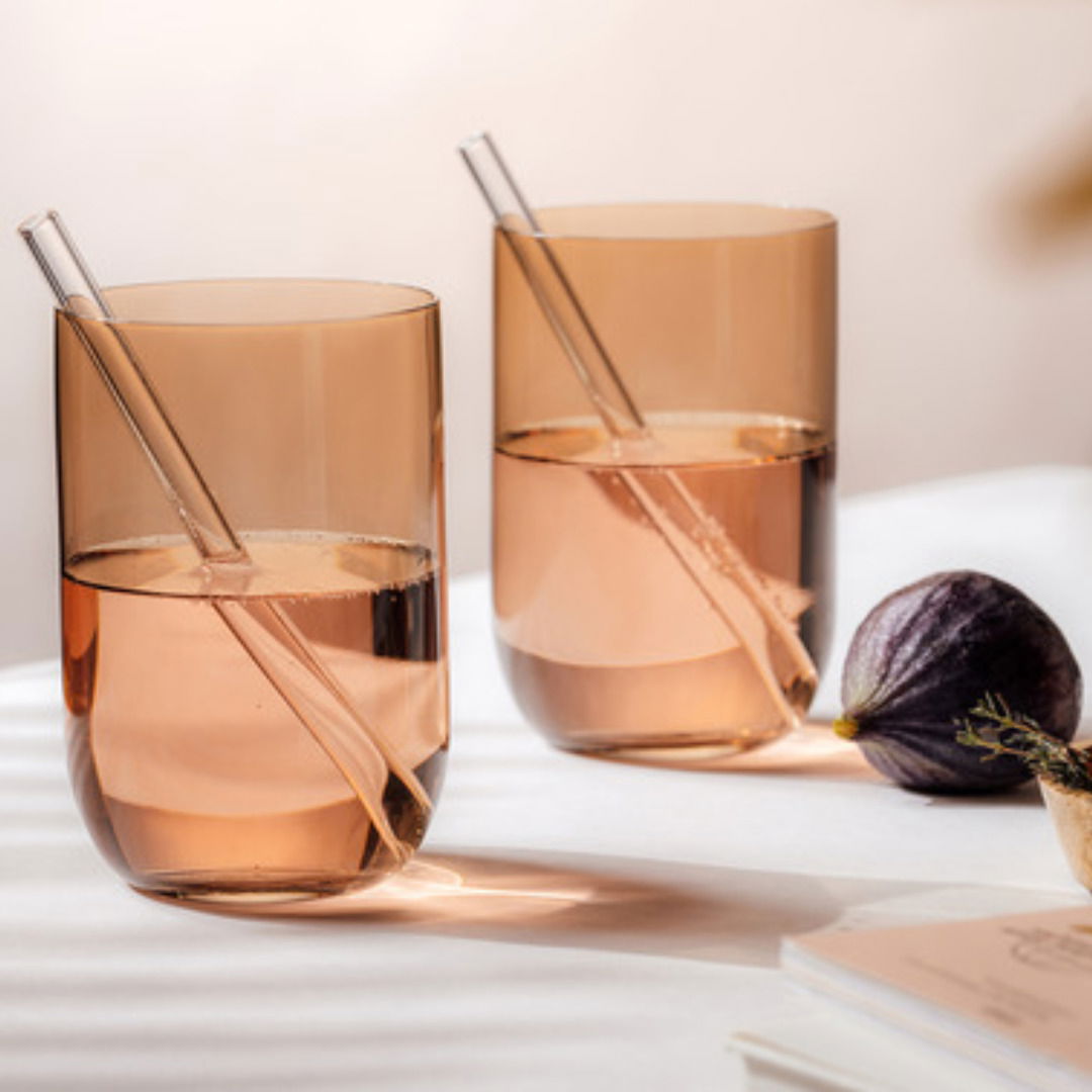 Zwei elegante Longdrinkbecher aus Glas mit bernsteinfarbenem Ton, jeweils mit einem Glasstrohhalm, auf einem Tisch mit weicher Beleuchtung und einer Feige im Vordergrund, seitliche Perspektive.