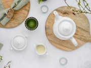 Draufsicht auf ein weißes Porzellan-Teeset mit Teekanne, Zuckerdose und Milchkännchen auf runden Holztabletts, daneben ein grünes Getränk im Glas sowie kleine Teelichter und Frühlingszweige.