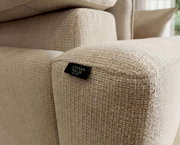 Detailaufnahme aus schräger Seitenperspektive eines beigen, grob strukturierten Polsterhockers/Sofateils mit schwarzem Markenlabel „JOHANN JAKOB“.