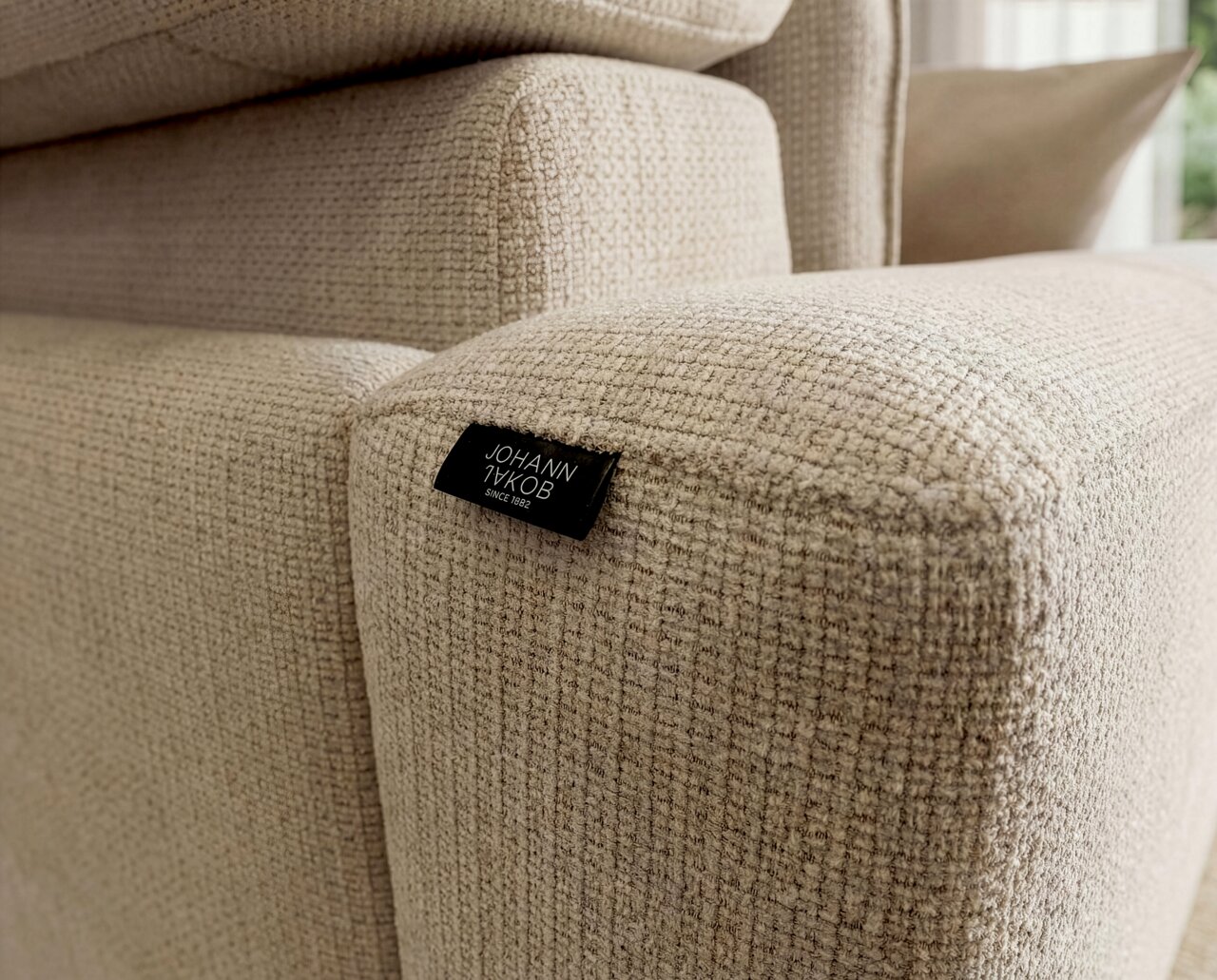 Detailaufnahme aus schräger Seitenperspektive eines beigen, grob strukturierten Polsterhockers/Sofateils mit schwarzem Markenlabel „JOHANN JAKOB“.