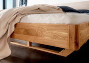 Massivholzbettgestell aus hellem Holz, seitliche Perspektive, mit Bettdecke und Kissen.