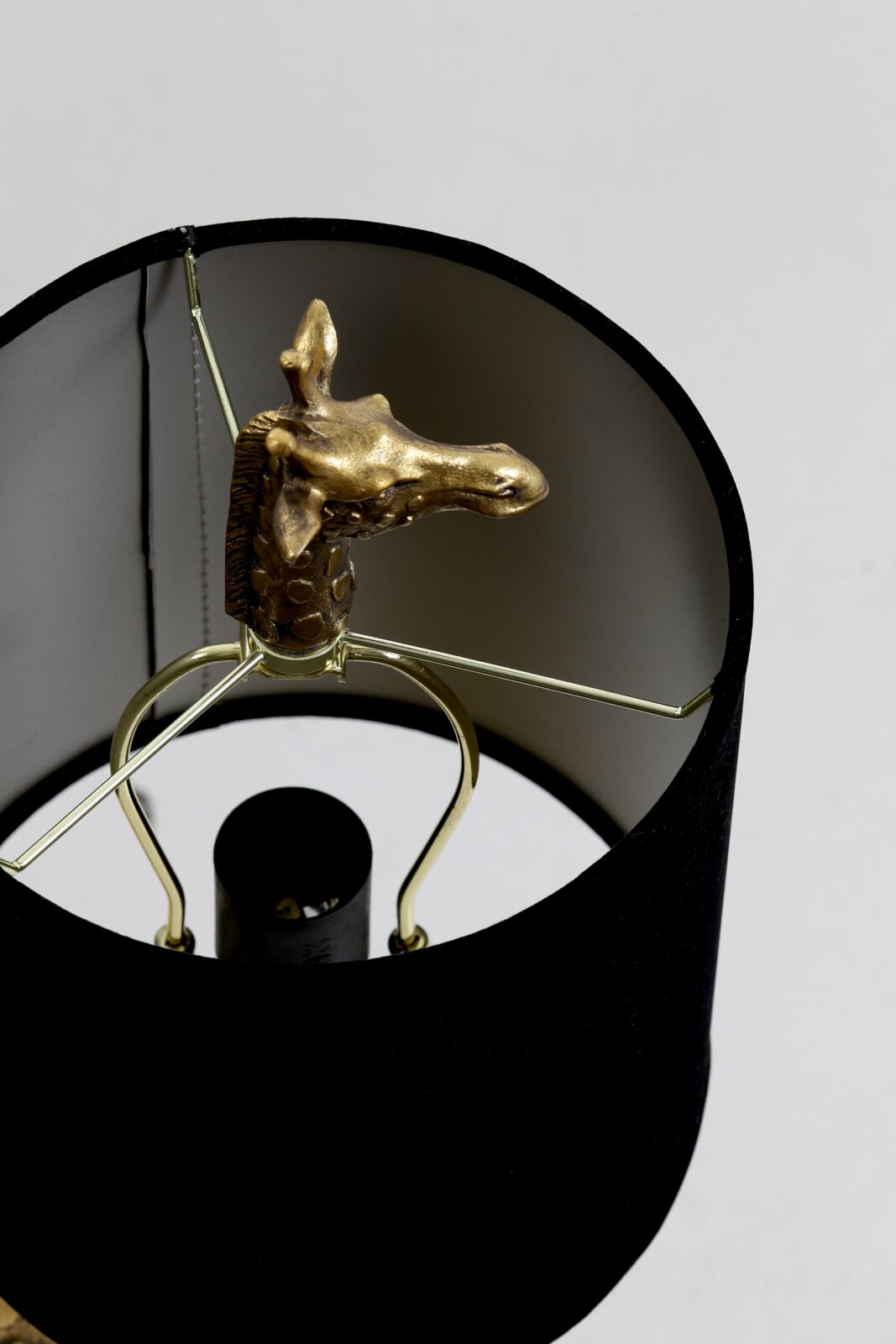 Close-up van een tafellamp met een zwarte lampenkap en een gouden giraffenfiguurtje erin, gezien van bovenaf.