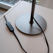 Runder Metallfuß einer schwarzen Tischlampe mit Kabel und Inline-Ein/Aus-Schalter auf weißer Tischplatte, schräg von oben fotografiert