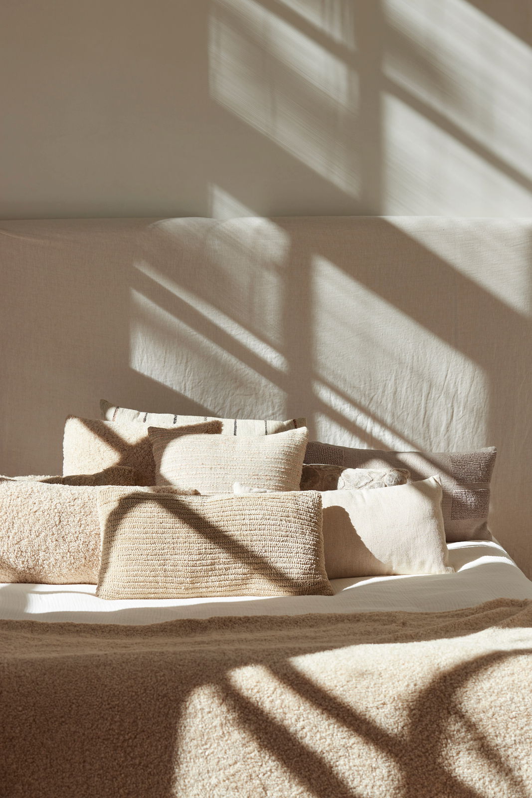 Beige Kissen in verschiedenen Größen auf einem Bett, beleuchtet von Sonnenlicht, das durch ein Fenster fällt. Perspektive von vorne.