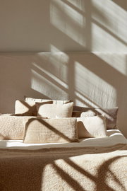 Beige Kissen in verschiedenen Größen auf einem Bett, beleuchtet von Sonnenlicht, das durch ein Fenster fällt. Perspektive von vorne.