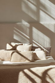 Beige Kissen in verschiedenen Größen auf einem Bett, beleuchtet von Sonnenlicht, das durch ein Fenster fällt. Perspektive von vorne.