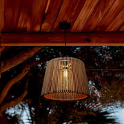 Hängelampe mit zylindrischem, vertikal geripptem Lampenschirm in Naturholz-Optik und sichtbarem Leuchtmittel, von unten leicht schräg aufgenommen unter einer Holzdecke im Außenbereich