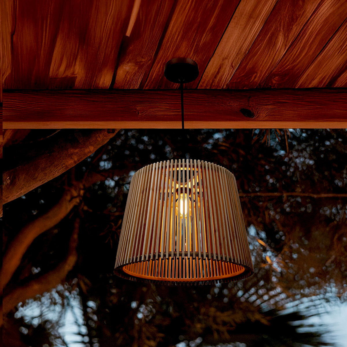 Newgarden Outdoor-Hängeleuchte OKINAWA Hängelampe mit zylindrischem, vertikal geripptem Lampenschirm in Naturholz-Optik und sichtbarem Leuchtmittel, von unten leicht schräg aufgenommen unter einer Holzdecke im Außenbereich