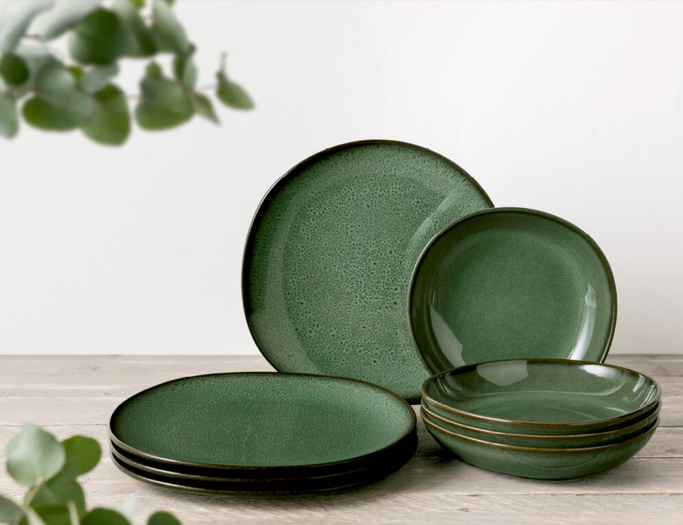 Grünes Keramik-Geschirrset, bestehend aus flachen und tiefen Tellern, auf einem Holztisch arrangiert. Seitliche Perspektive.