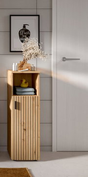 Schmaler Badschrank aus hellem Holz mit vertikalen Lamellenfronten, offenem Ablagefach und schwarzem Griff, frontale Perspektive im Badezimmer mit Deko auf der Ablage