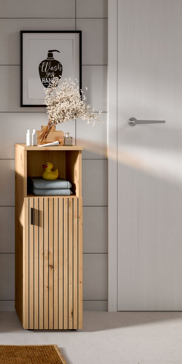 Schmaler Badschrank aus hellem Holz mit vertikalen Lamellenfronten, offenem Ablagefach und schwarzem Griff, frontale Perspektive im Badezimmer mit Deko auf der Ablage