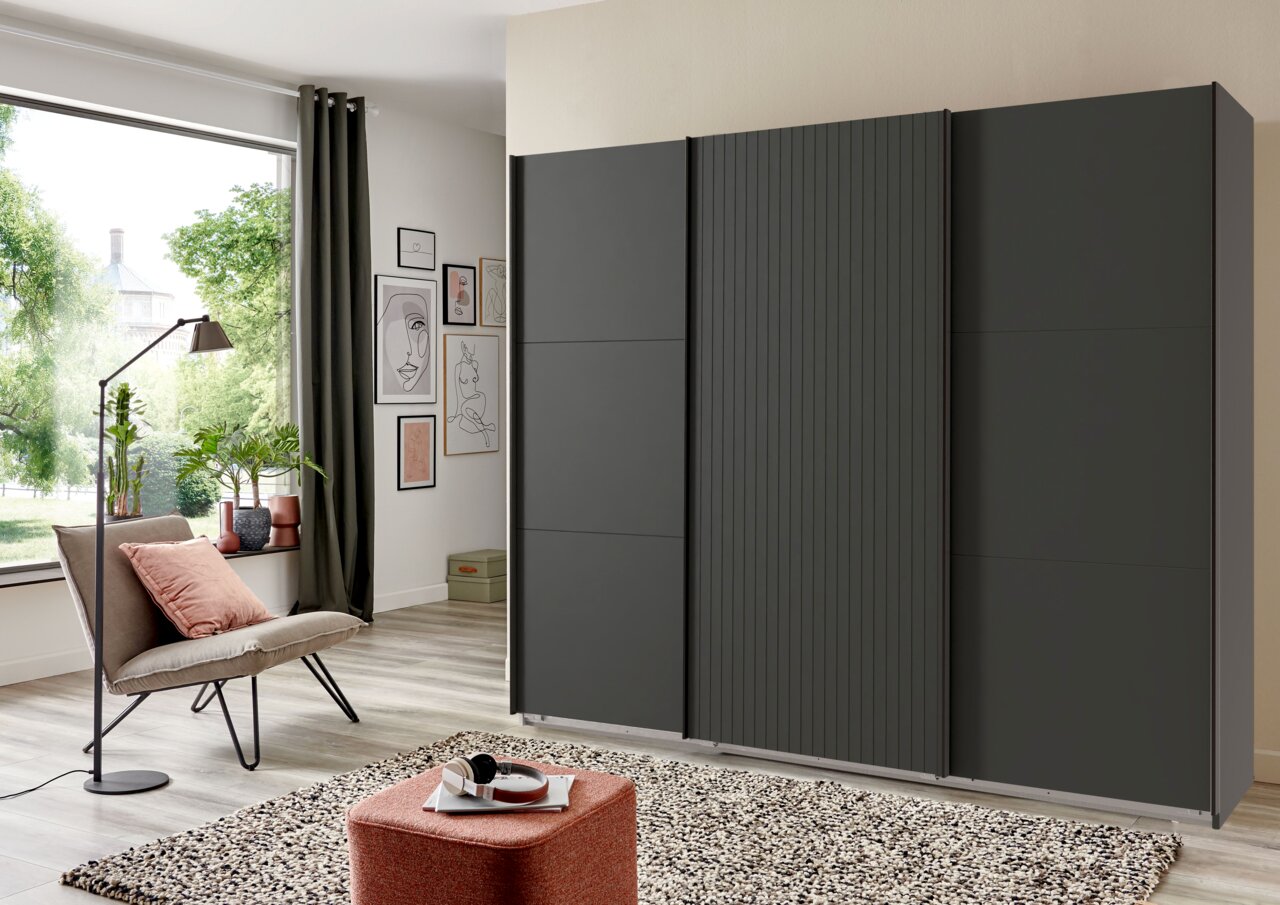 Dreitüriger Schwebetürenschrank in dunklem Design, frontal fotografiert in einem modernen Wohnzimmer mit großen Fenstern und stilvoller Dekoration.