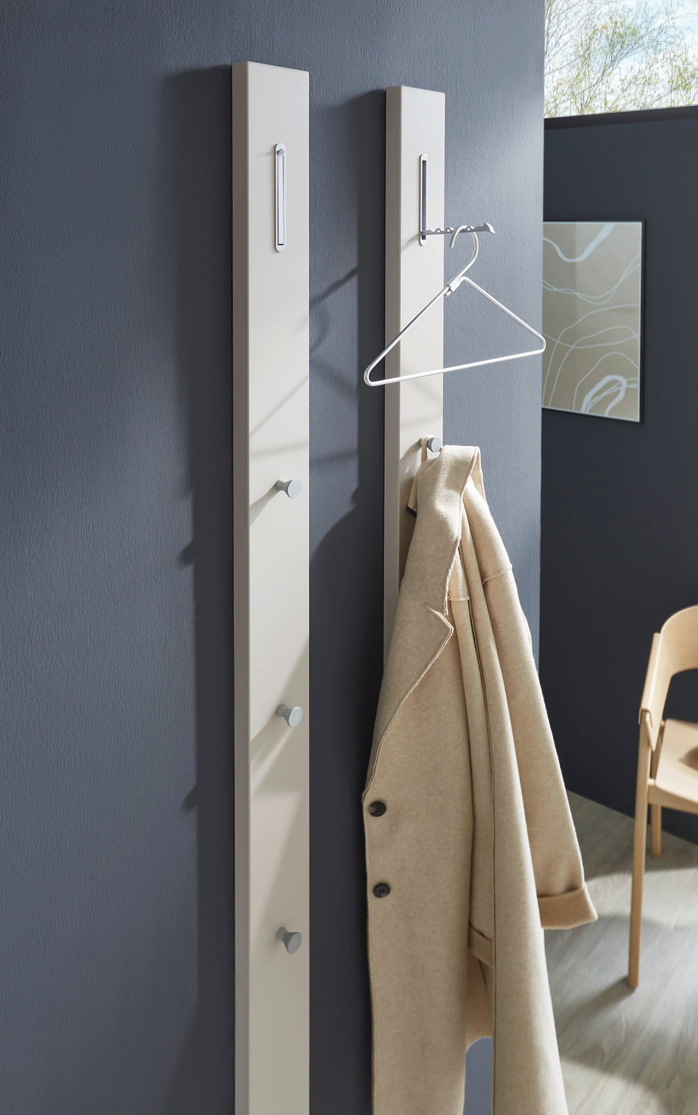 Zwei moderne Garderobenpaneele in Beige an einer dunkelgrauen Wand, mit Haken und einem beigen Mantel, aus seitlicher Perspektive fotografiert.