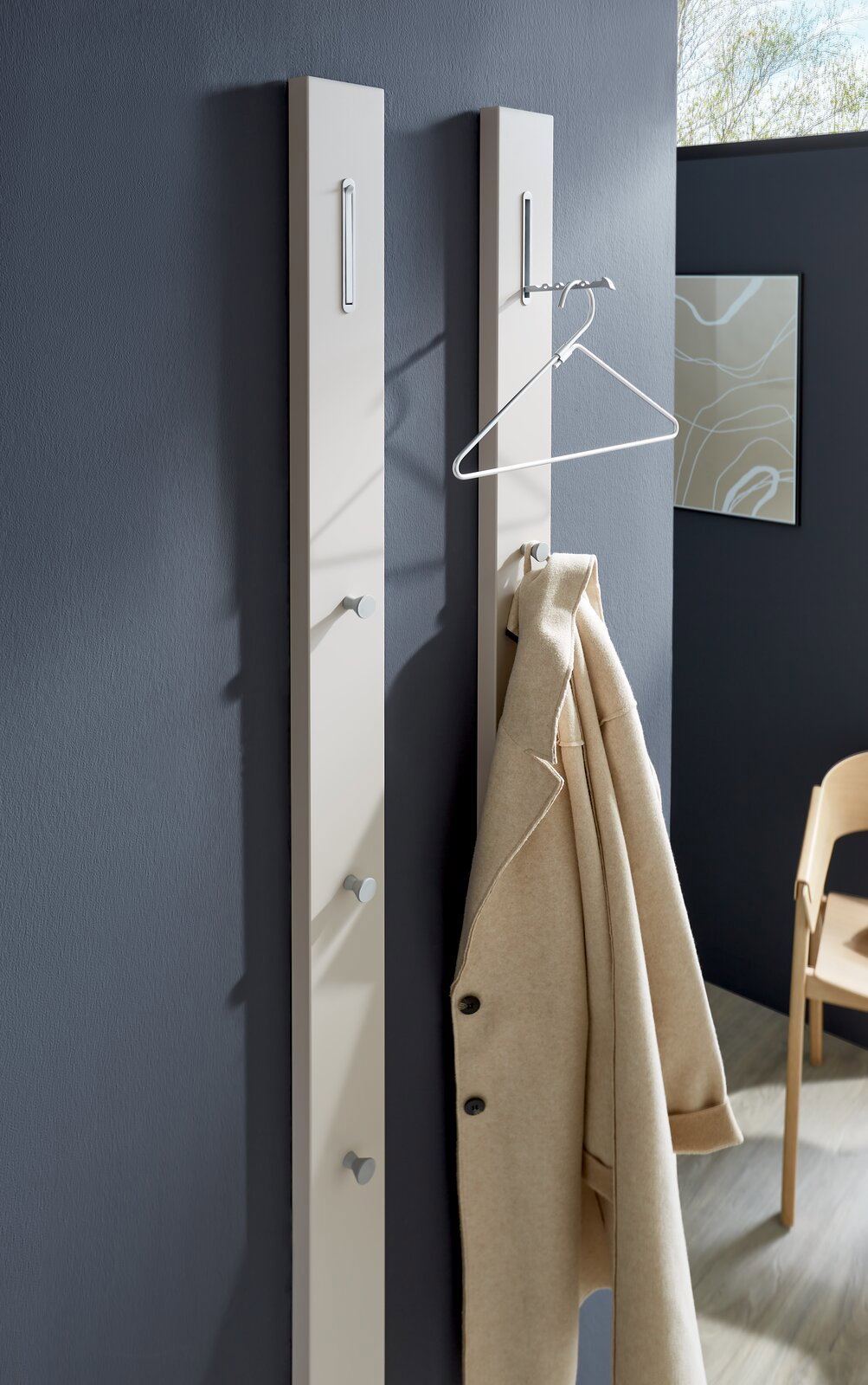 Zwei moderne Garderobenpaneele in Beige an einer dunkelgrauen Wand, mit Haken und einem beigen Mantel, aus seitlicher Perspektive fotografiert.