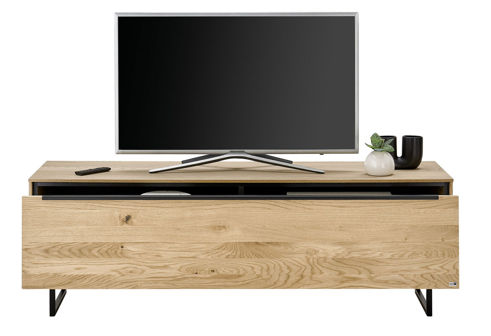 TV-Lowboard klein aus Holz mit schwarzem Metallgestell, darauf ein Flachbildfernseher, Fernbedienung und Dekoration. Frontalansicht.