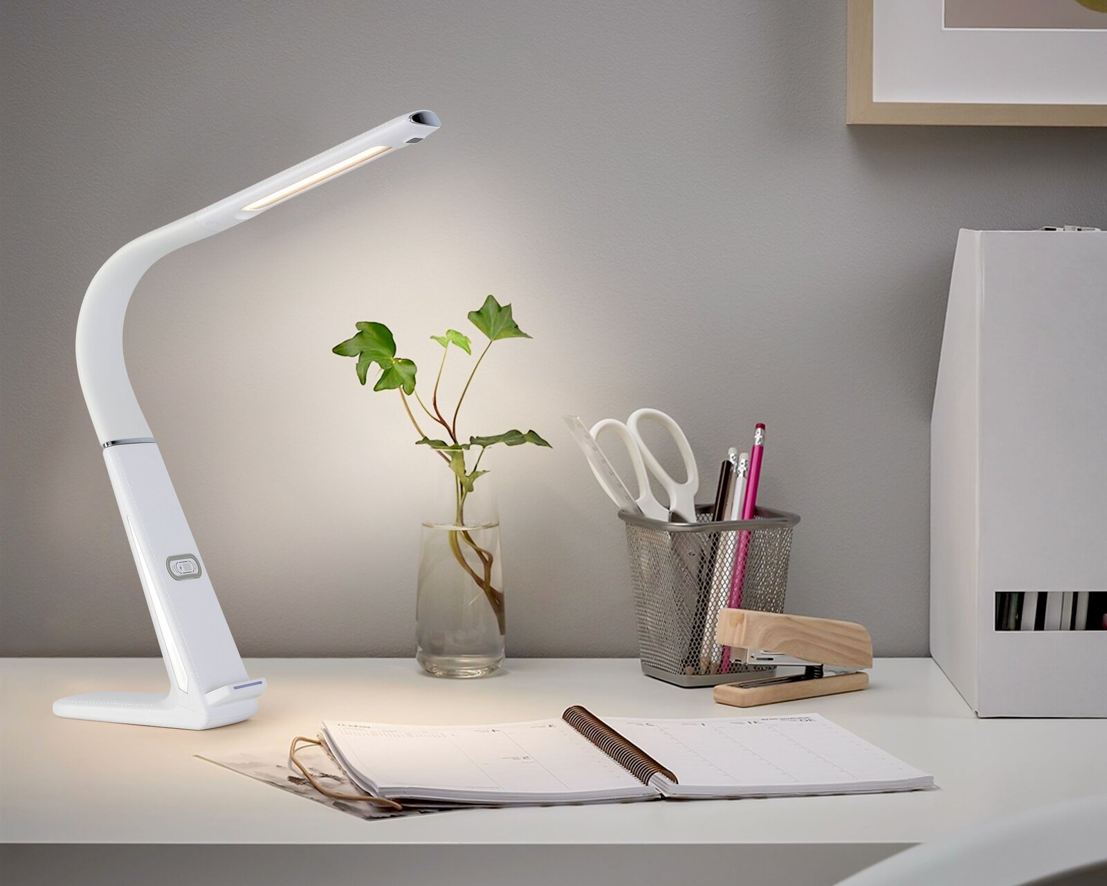 Moderne LED-Schreibtischleuchte in Weiß, seitliche Perspektive, auf einem Schreibtisch neben Büromaterialien und einer Pflanze.