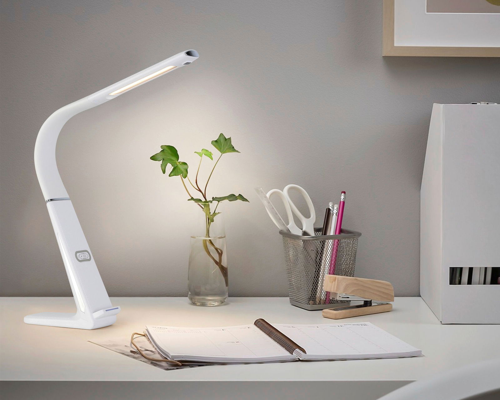 Moderne LED-Schreibtischleuchte in Weiß, seitliche Perspektive, auf einem Schreibtisch neben Büromaterialien und einer Pflanze.
