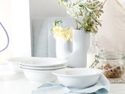 Weiße Keramikschalen und Teller auf einem Tisch neben Glas mit Müsli und Milchflasche, im Hintergrund eine geriffelte weiße Vase mit Blumen; Perspektive: leicht seitliche Nahaufnahme auf Tischhöhe.