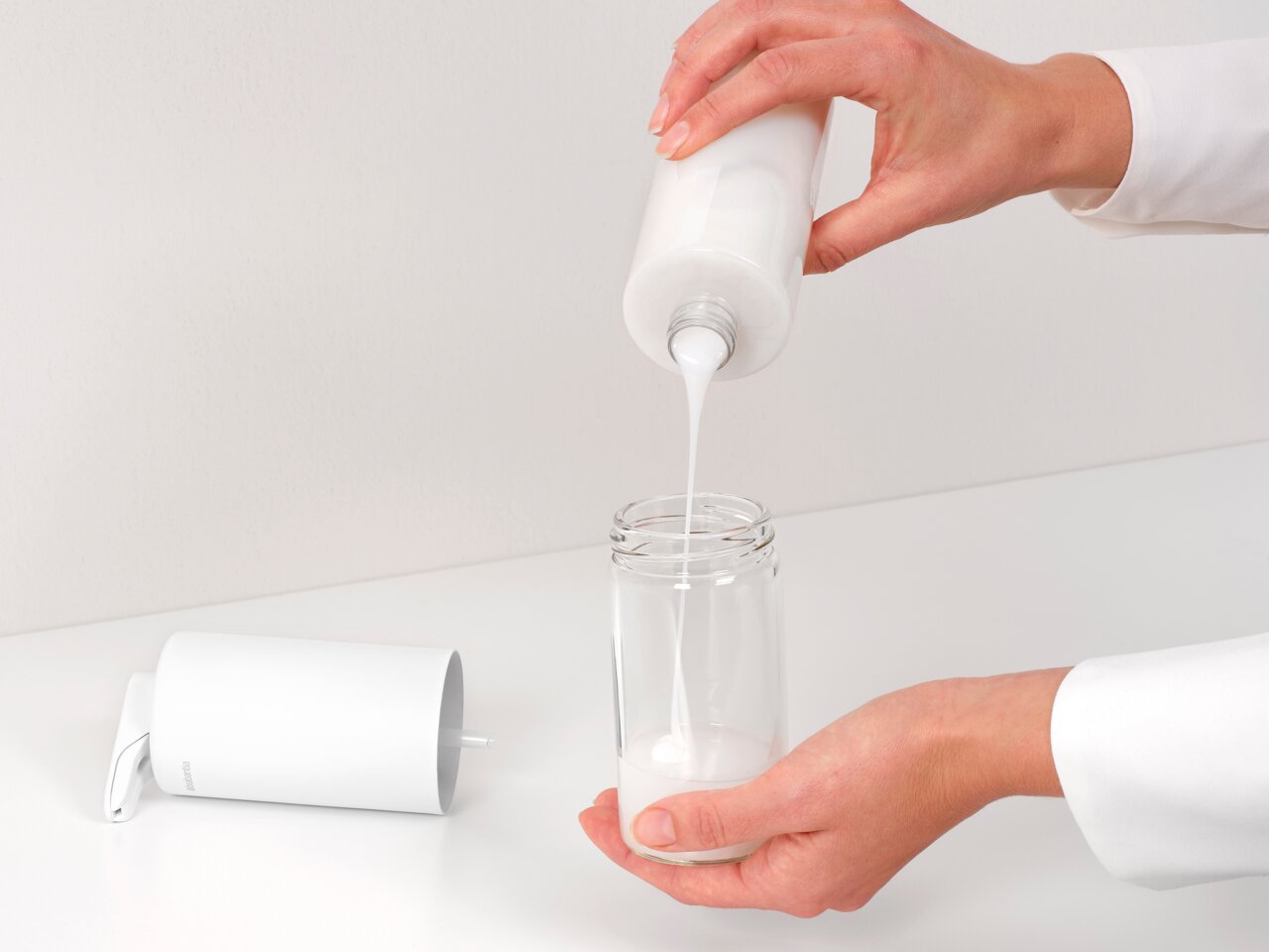 Close-up van een Brabantia SINKSTYLE-zeepdispenser in wit, die op een wit oppervlak staat, terwijl een hand vloeibare zeep in een doorzichtig bakje giet.