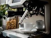 Nahaufnahme einer Edelstahl-Siebträger-Espressomaschine mit Dampflanze und eingespanntem Siebträger, vorne eine weiße Espressotasse auf der Abtropfschale; Perspektive: seitlich von vorn auf Höhe der Brühgruppe