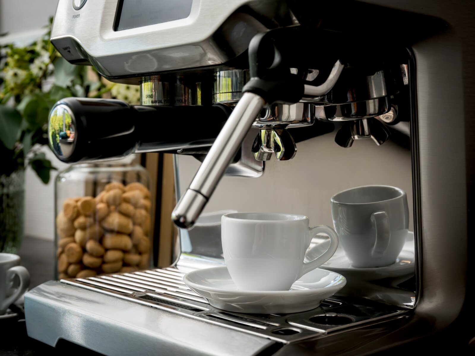 Nahaufnahme einer Edelstahl-Siebträger-Espressomaschine mit Dampflanze und eingespanntem Siebträger, vorne eine weiße Espressotasse auf der Abtropfschale; Perspektive: seitlich von vorn auf Höhe der Brühgruppe