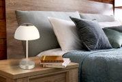 Moderne Tischleuchte mit weißem Schirm auf einem Nachttisch, neben einem Bett mit grauen und grünen Kissen, aus seitlicher Perspektive.