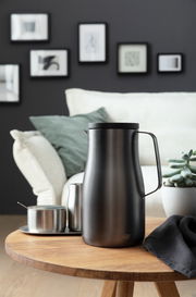 Elegante Isolierkanne Studio in mattem Schwarz auf einem Holztisch, umgeben von modernen Wohnaccessoires, seitliche Perspektive.