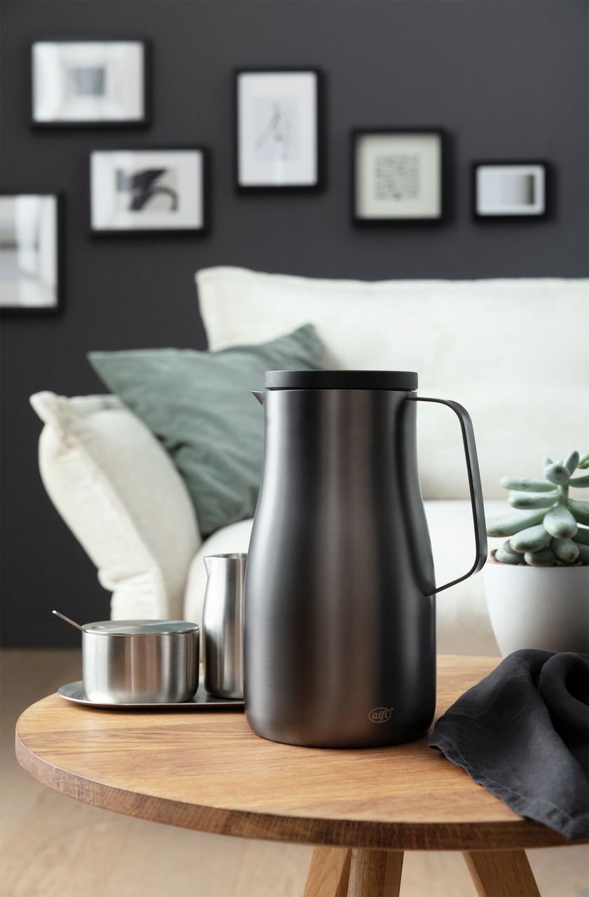 Elegante Isolierkanne Studio in mattem Schwarz auf einem Holztisch, umgeben von modernen Wohnaccessoires, seitliche Perspektive.
