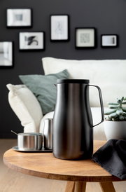 Elegante Isolierkanne Studio in mattem Schwarz auf einem Holztisch, umgeben von modernen Wohnaccessoires, seitliche Perspektive.
