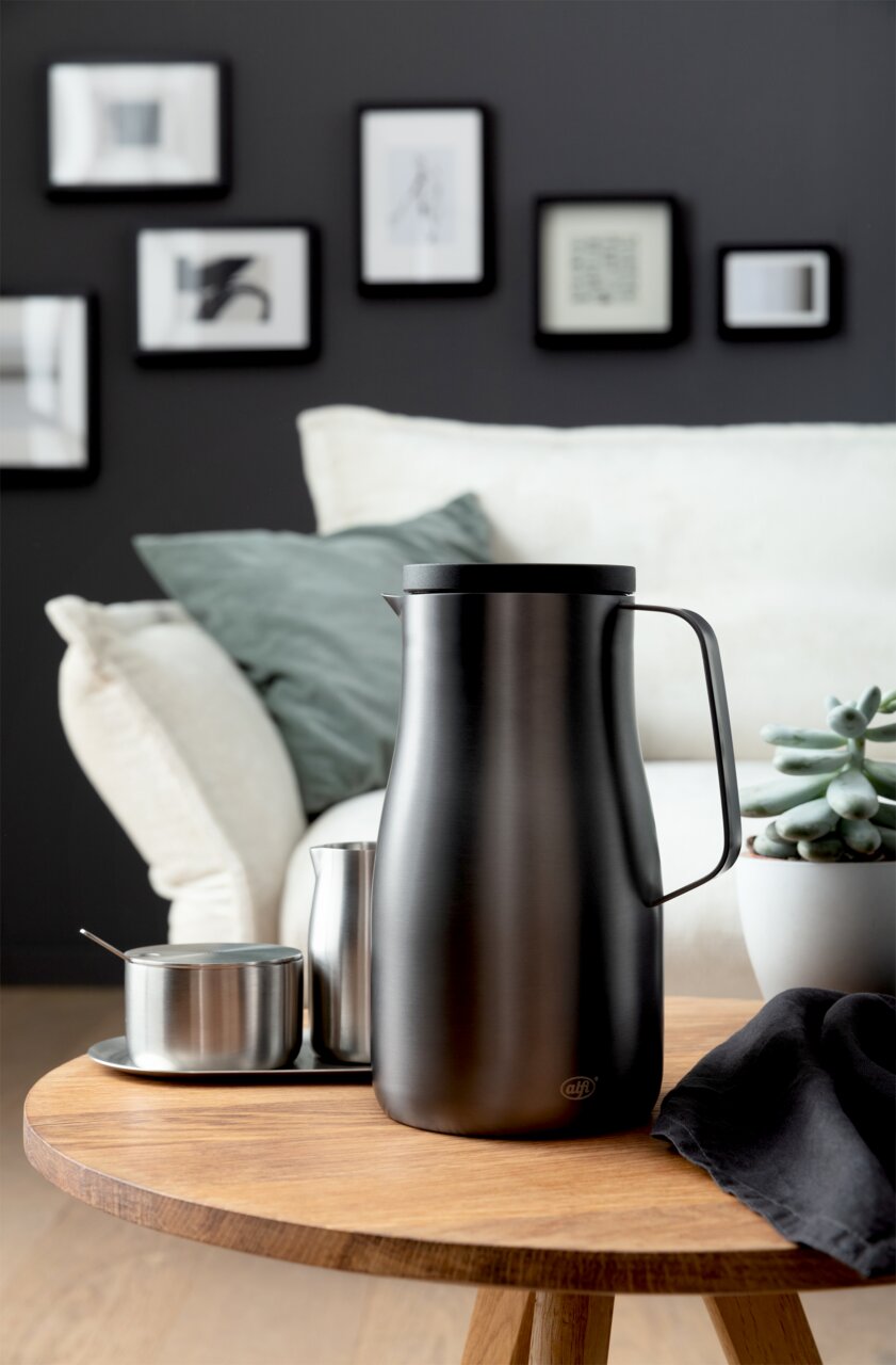 Elegante Isolierkanne Studio in mattem Schwarz auf einem Holztisch, umgeben von modernen Wohnaccessoires, seitliche Perspektive.
