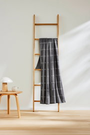 Grijze strandlaken van 90 x 180 cm met ruitjespatroon, gedrapeerd over een houten ladder in een minimalistische ruimte, gezien vanuit een zijdelingse hoek.