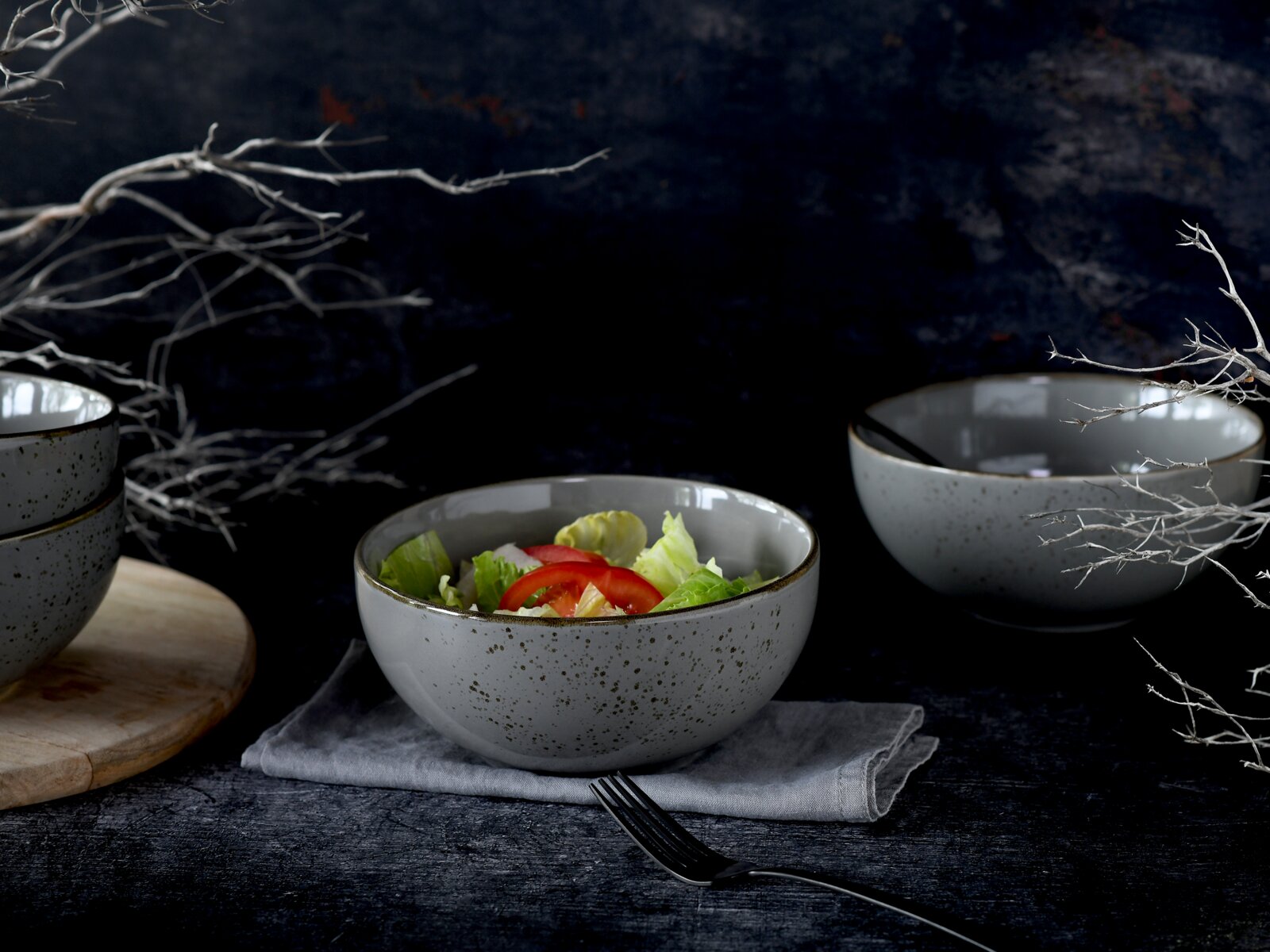 Grau gesprenkelte Keramik-Schüssel mit Salat und Tomatenscheiben auf Leinentuch, in leicht seitlicher Perspektive auf dunklem Tisch, weitere Schalen im Hintergrund