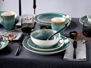 Modern gedeckter Tisch mit blau-grün glasiertem Keramikgeschirr (Teller, Schale, Becher), schwarzen Besteckteilen und geriffeltem Glas, dekoriert mit roten Beerenzweigen, fotografiert aus leicht erhöhter schräger Vorderansicht.