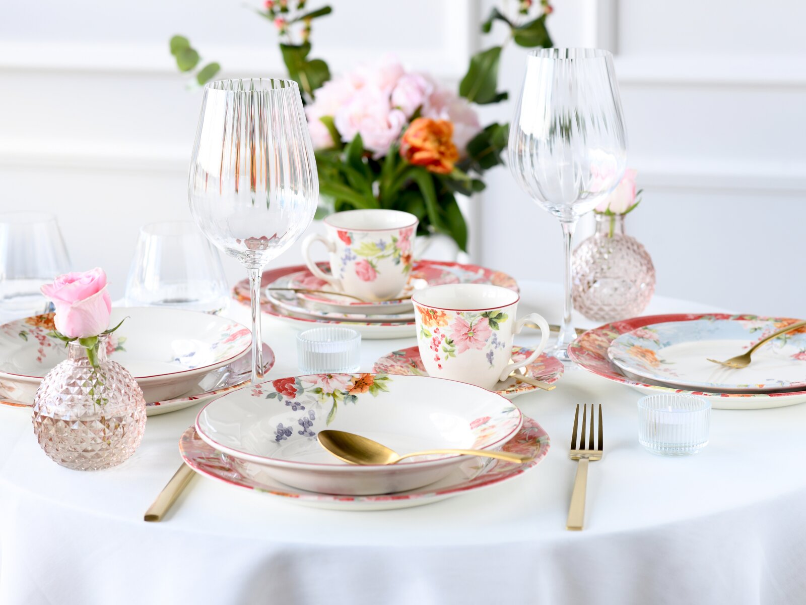 Frühlingshaft gedeckter Tisch mit floralem Porzellangeschirr, zwei großen geriffelten Weingläsern, goldfarbenem Besteck und rosa Glasvasen mit Rosen, aufgenommen in leichter Schrägansicht von vorn.