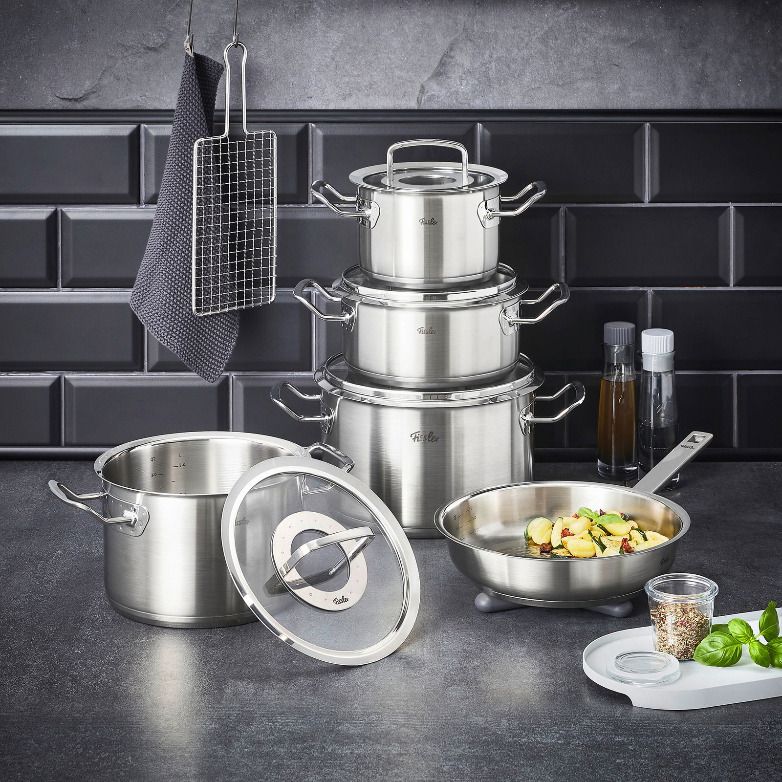 Topfset Original-Profi 5-tlg. aus Edelstahl, bestehend aus mehreren Töpfen und einer Pfanne, auf einer Küchenarbeitsplatte vor einer dunklen Fliesenwand, mit Küchenutensilien im Hintergrund.
