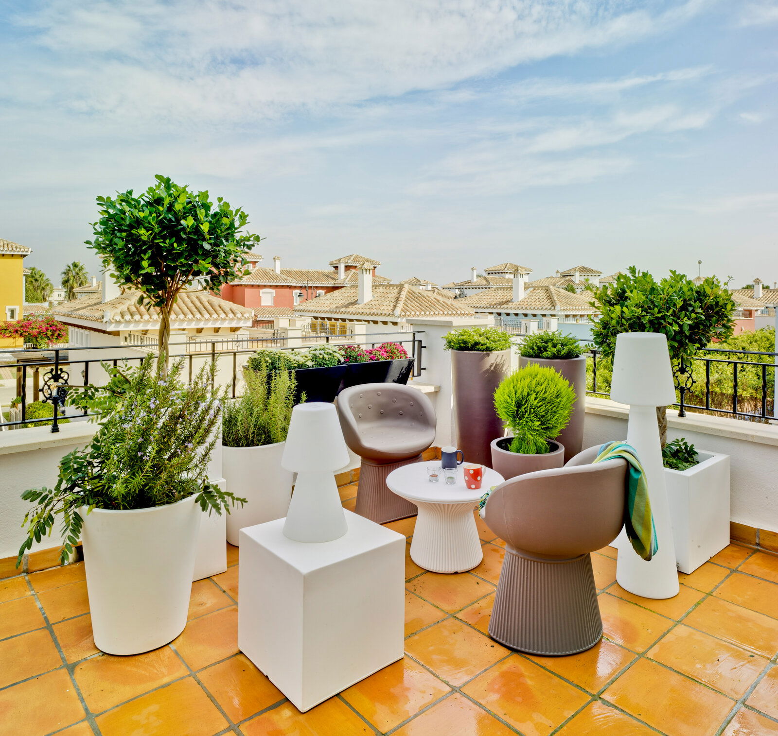 Moderne Dachterrasse mit zwei taupefarbenen Lounge-Sesseln, rundem weißem Beistelltisch, weißen Outdoor-Leuchten und großen Pflanzkübeln, Blick aus leicht erhöhter Perspektive über die Sitzgruppe auf mediterrane Hausdächer und blauen Himmel