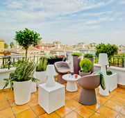 Moderne Dachterrasse mit zwei taupefarbenen Lounge-Sesseln, rundem weißem Beistelltisch, weißen Outdoor-Leuchten und großen Pflanzkübeln, Blick aus leicht erhöhter Perspektive über die Sitzgruppe auf mediterrane Hausdächer und blauen Himmel