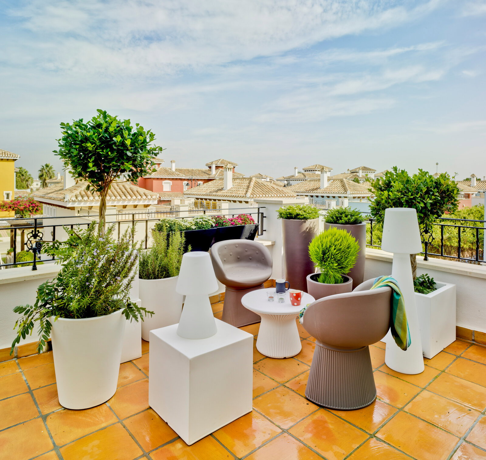 Moderne Dachterrasse mit zwei taupefarbenen Lounge-Sesseln, rundem weißem Beistelltisch, weißen Outdoor-Leuchten und großen Pflanzkübeln, Blick aus leicht erhöhter Perspektive über die Sitzgruppe auf mediterrane Hausdächer und blauen Himmel