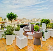 Moderne Dachterrasse mit zwei taupefarbenen Lounge-Sesseln, rundem weißem Beistelltisch, weißen Outdoor-Leuchten und großen Pflanzkübeln, Blick aus leicht erhöhter Perspektive über die Sitzgruppe auf mediterrane Hausdächer und blauen Himmel