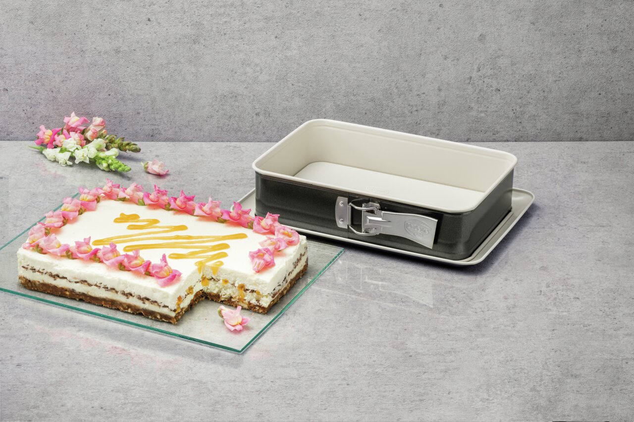 Rechteckige Backform mit abnehmbarem Rand, präsentiert neben einem dekorierten Kuchen mit Blumen und Fruchtsoße. Perspektive von oben.