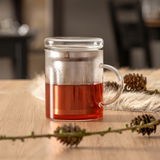 Teeglas mit Filter und Deckel, gefüllt mit rotem Tee, auf einem Holztisch mit Kiefernzapfen, seitliche Perspektive