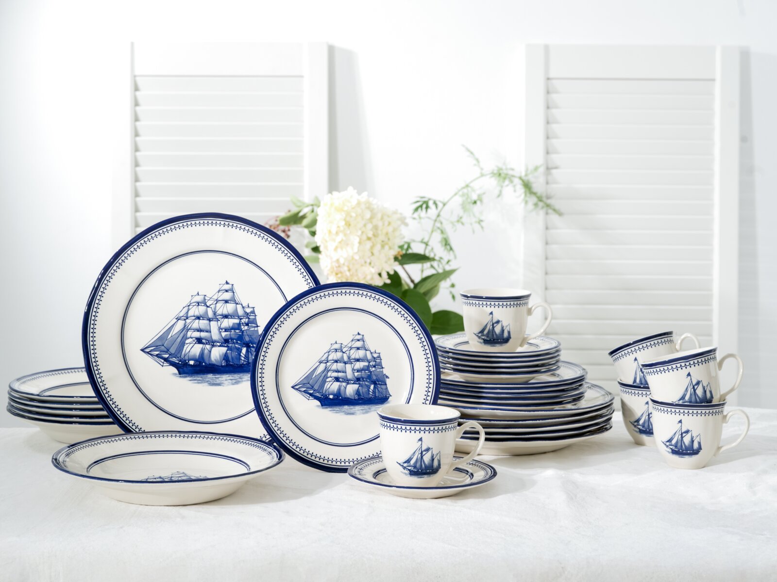 Maritimes blau-weißes Porzellan-Geschirrset mit Segelschiff-Motiv, Teller, Schalen und Tassen auf weißem Tisch arrangiert, fotografiert in frontaler leicht seitlicher Perspektive.