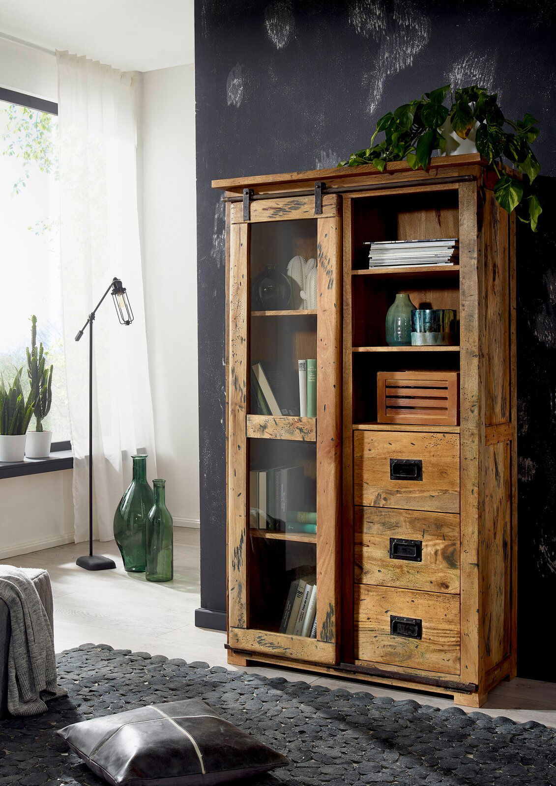 Holzvitrine mit Schiebetür und Regalen, gefüllt mit Büchern und Dekorationen, aus der Perspektive eines Wohnzimmers mit Pflanzen und Stehlampe.