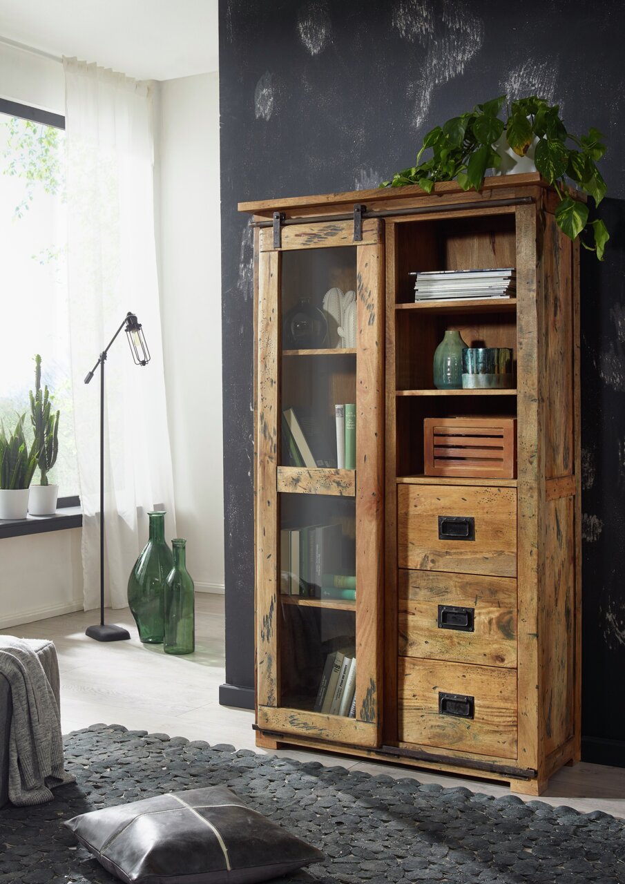 Holzvitrine mit Schiebetür und Regalen, gefüllt mit Büchern und Dekorationen, aus der Perspektive eines Wohnzimmers mit Pflanzen und Stehlampe.