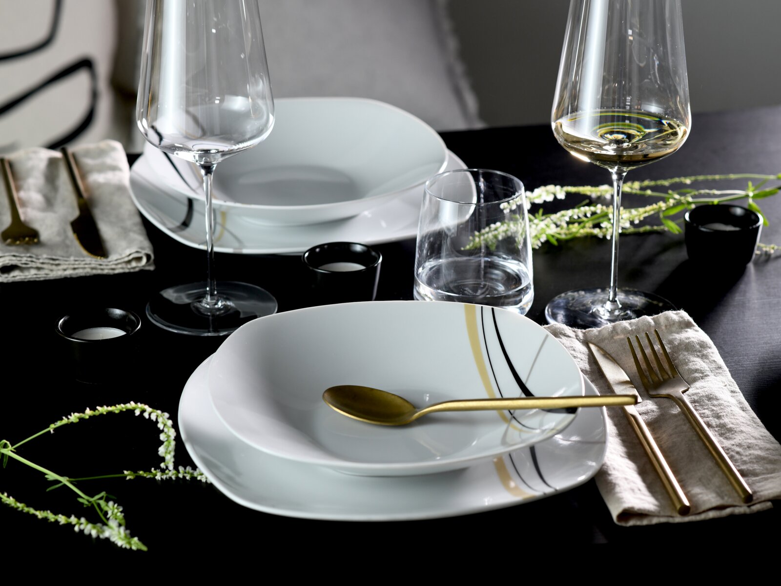 Schwarz gedeckter Tisch mit weißem Porzellan-Geschirr mit gold-schwarzen Linien, goldfarbenem Besteck, Weingläsern und Wasserglas, aufgenommen in schräger Draufsicht.