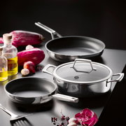 Edelstahl-Kochgeschirr-Set mit Pfanne, flacher Kasserolle mit Glasdeckel und weiterer Pfanne auf dunkler Arbeitsfläche, umgeben von Gemüse und Ölflaschen, fotografiert aus leicht erhöhter schräger Perspektive