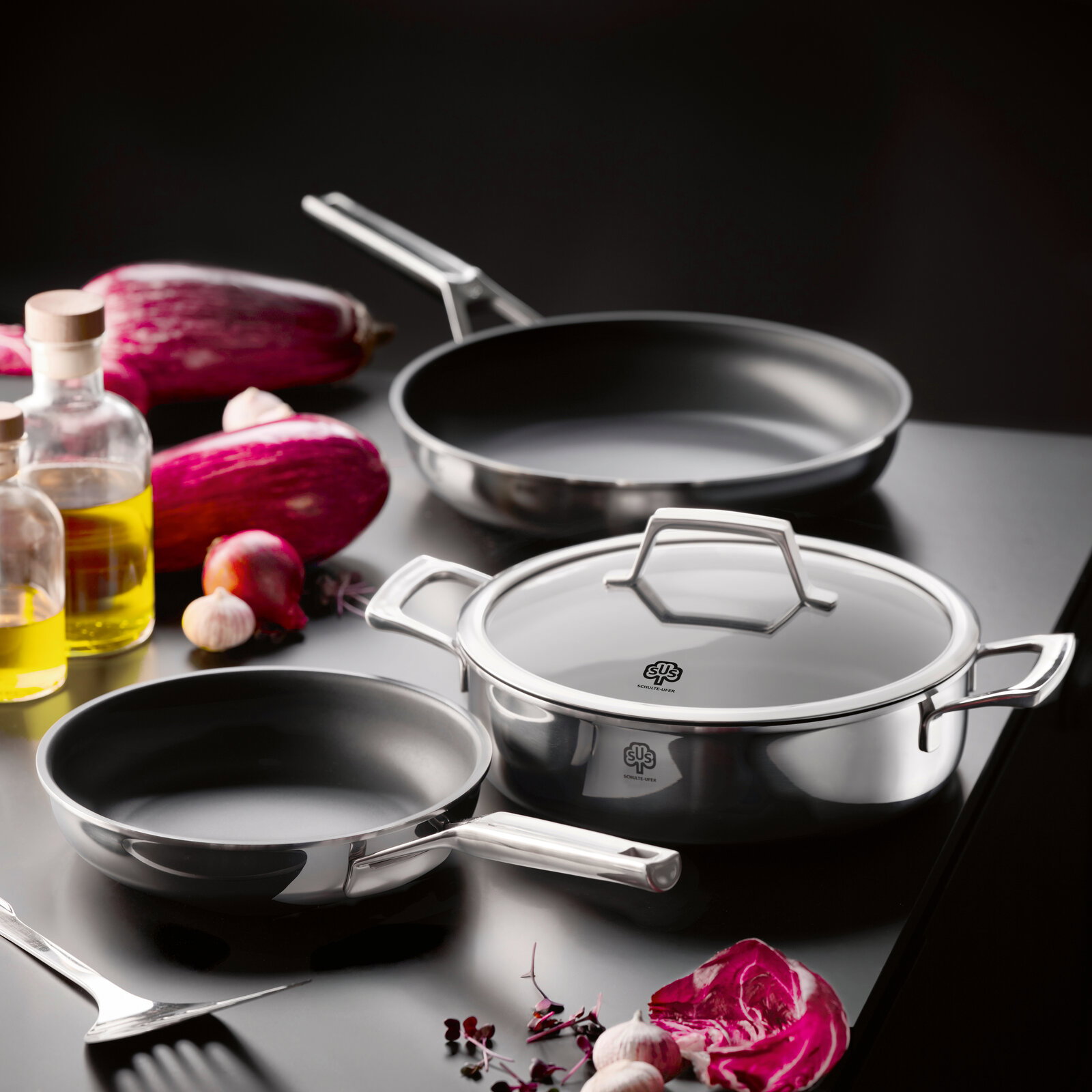 Edelstahl-Kochgeschirr-Set mit Pfanne, flacher Kasserolle mit Glasdeckel und weiterer Pfanne auf dunkler Arbeitsfläche, umgeben von Gemüse und Ölflaschen, fotografiert aus leicht erhöhter schräger Perspektive