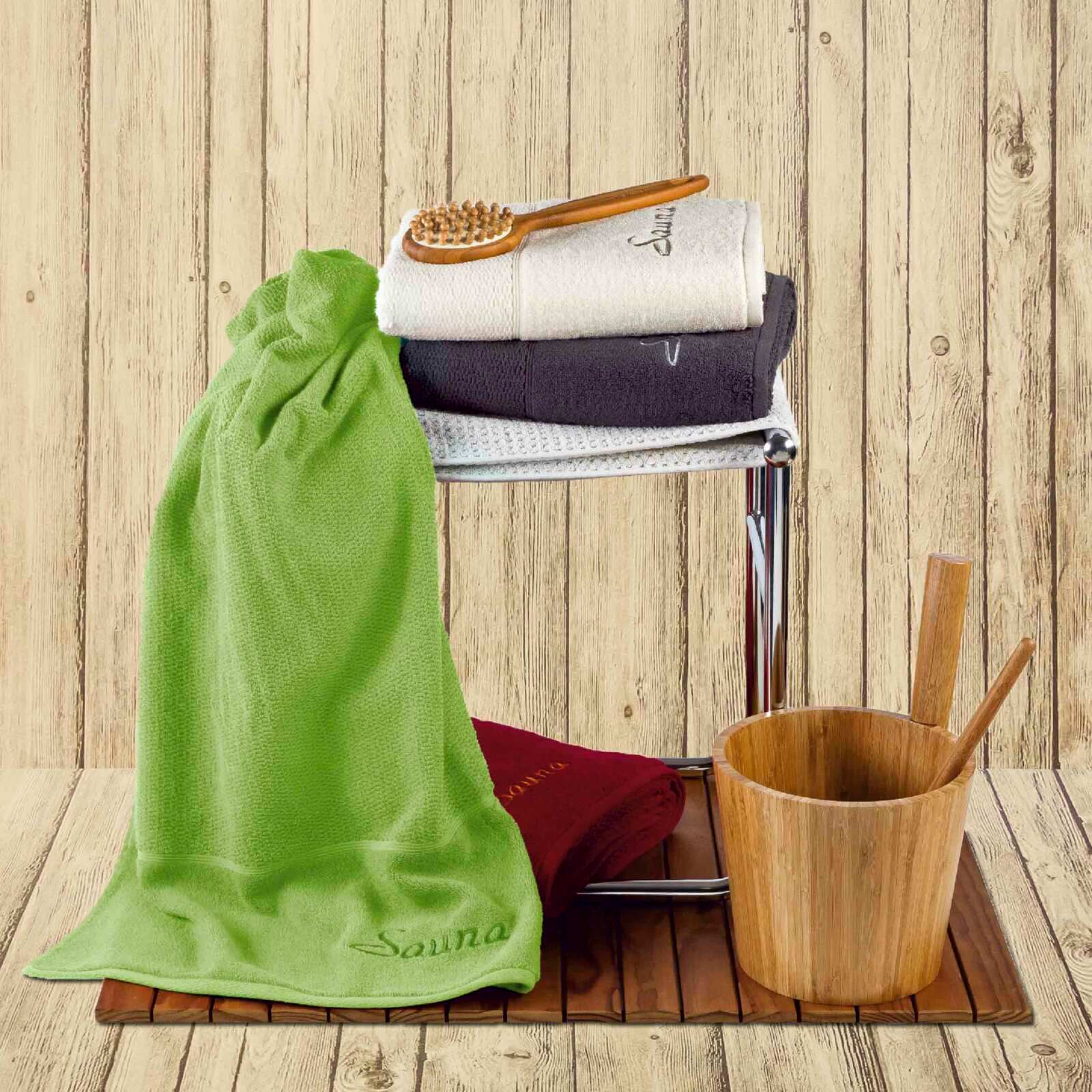 MÖVE Saunatuch 2er-Set PEARL Saunatuch-Set in verschiedenen Farben, gestapelt auf einem Metallregal mit Holzgriffbürste, vor einer Holzwand. Perspektive: Frontalansicht.